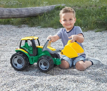 Ein lächelnder Junge kniet auf Kies und spielt mit einem grünen und gelben GIGA TRUCKS Traktor mit Frontlader, dessen Schaufel mit Steinen gefüllt ist.
