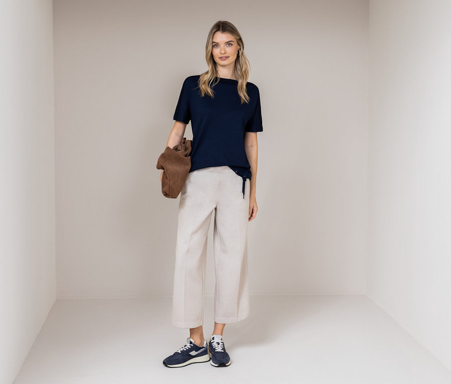 Eine Frau steht in einem Raum und trägt ein dunkelblaues T-Shirt, beigefarbene Hosen und blaue Turnschuhe. Sie hält eine braune Tasche.