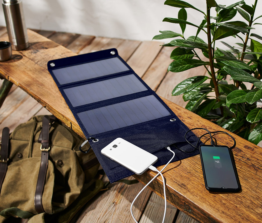 Ein faltbares Solar-Ladegerät lädt zwei Telefone auf einer Holzbank neben einem Rucksack auf.