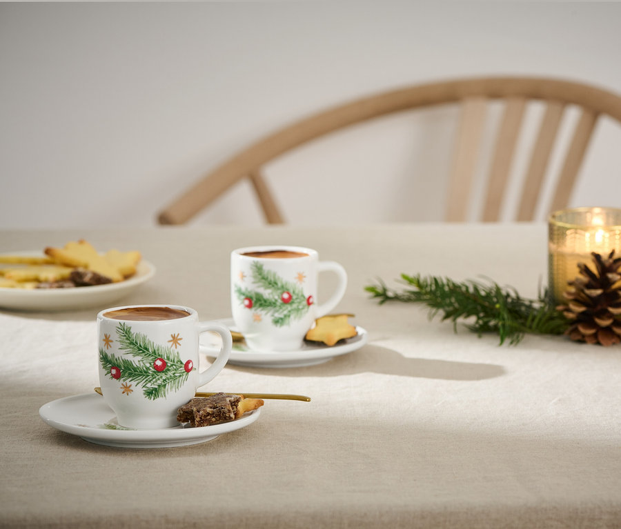 Zwei Espressotassen mit Weihnachtsmotiv auf einem Tisch mit Keksen und Kerzen.