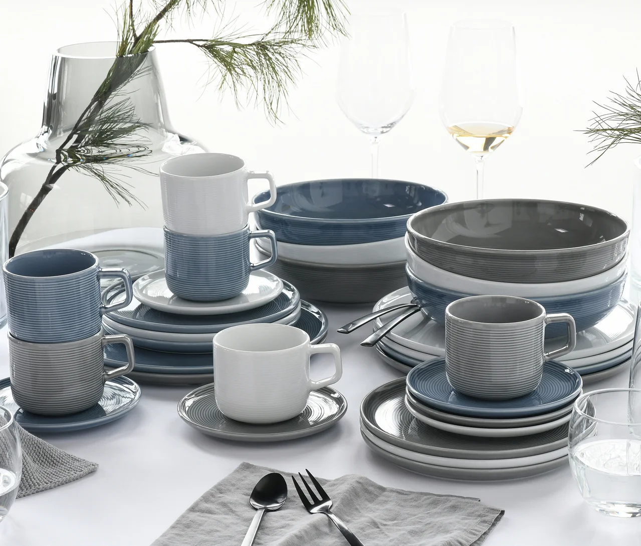 Ein gedeckter Tisch mit dem creaTable Service »Session Urban«, 30-teilig, bestehend aus gerippten Tassen, Untertassen, Tellern und Schüsseln in Weiß, Blau und Grau. Das Geschirr ist auf einem weißen Tischtuch arrangiert, daneben Weingläser, Besteck auf grauen Servietten und eine Glasvase mit Kiefernzweigen.