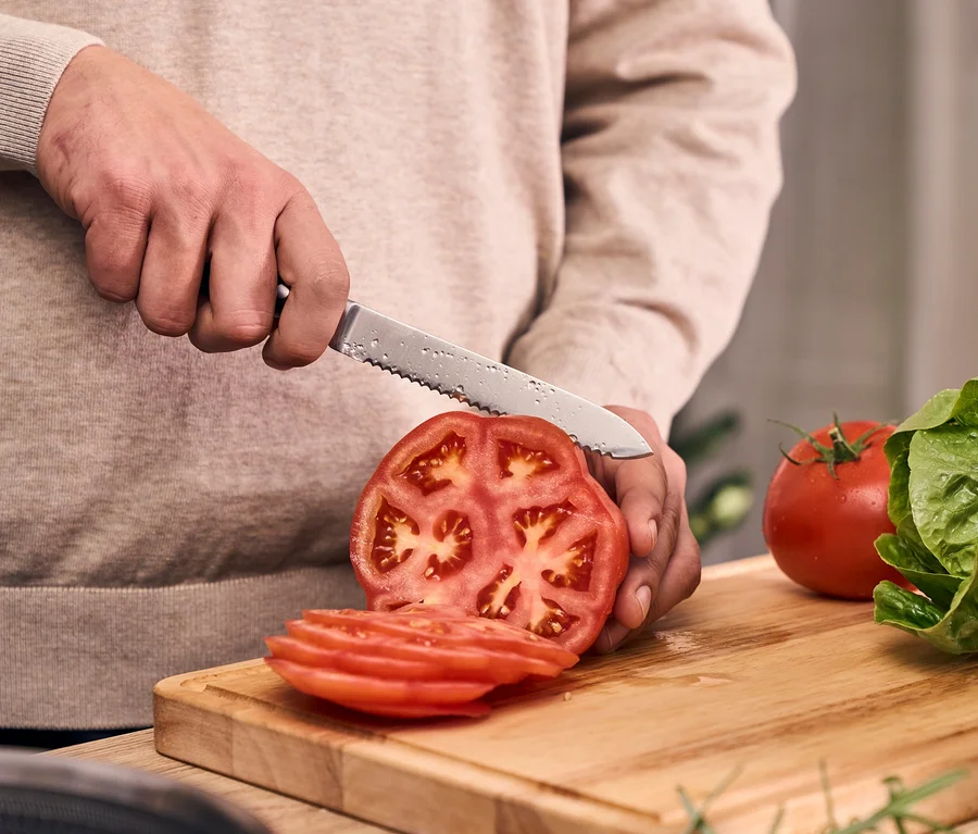 Hände schneiden eine rote Tomate mit einem gezackten Messer aus dem Henckels by ZWILLING »Pakka« Messerblockset auf einem Holzschneidebrett. Daneben liegen Tomatenscheiben, eine ganze Tomate und grüner Salat.