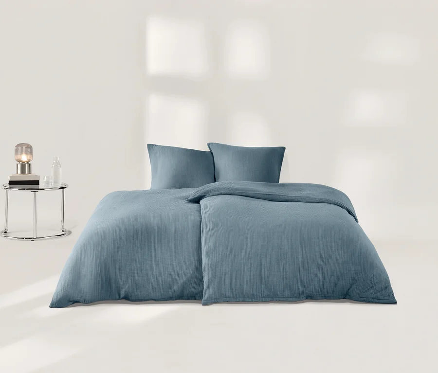 Blaue Musselin-Bettwäsche mit zwei Kissen und zwei Bettbezügen auf weißer Fläche. Links ein Nachttisch mit geriffelter Lampe, Glasflasche und Glas.