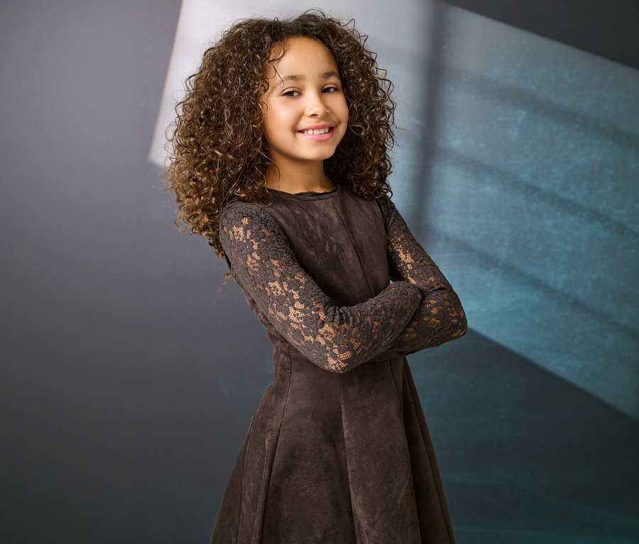 Lächelndes junges Mädchen mit lockigem braunem Haar trägt ein braunes Kleid mit Spitzärmeln und verschränkten Armen vor dunklem Hintergrund.