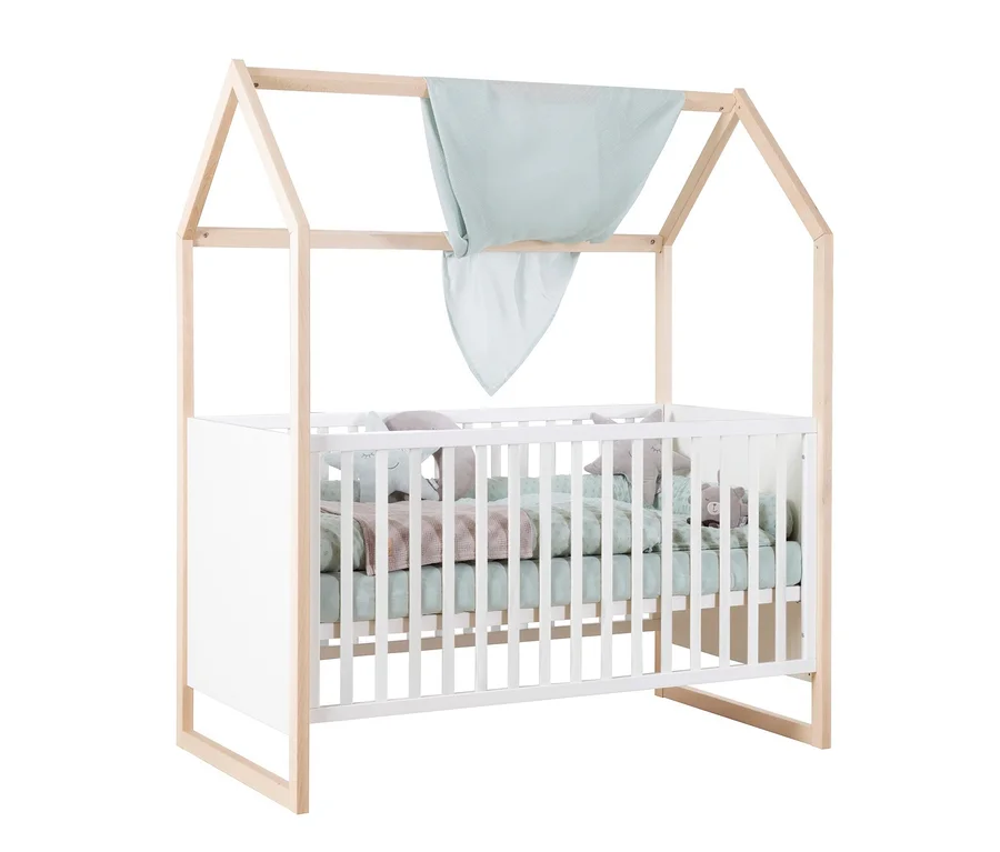 Weißes Babybett mit einem hellen Holzrahmen in Hausform, über dem ein hellblaues Tuch hängt. Im Bett liegen hellgrüne Bettwäsche, eine rosa Decke und mehrere Stofftiere.