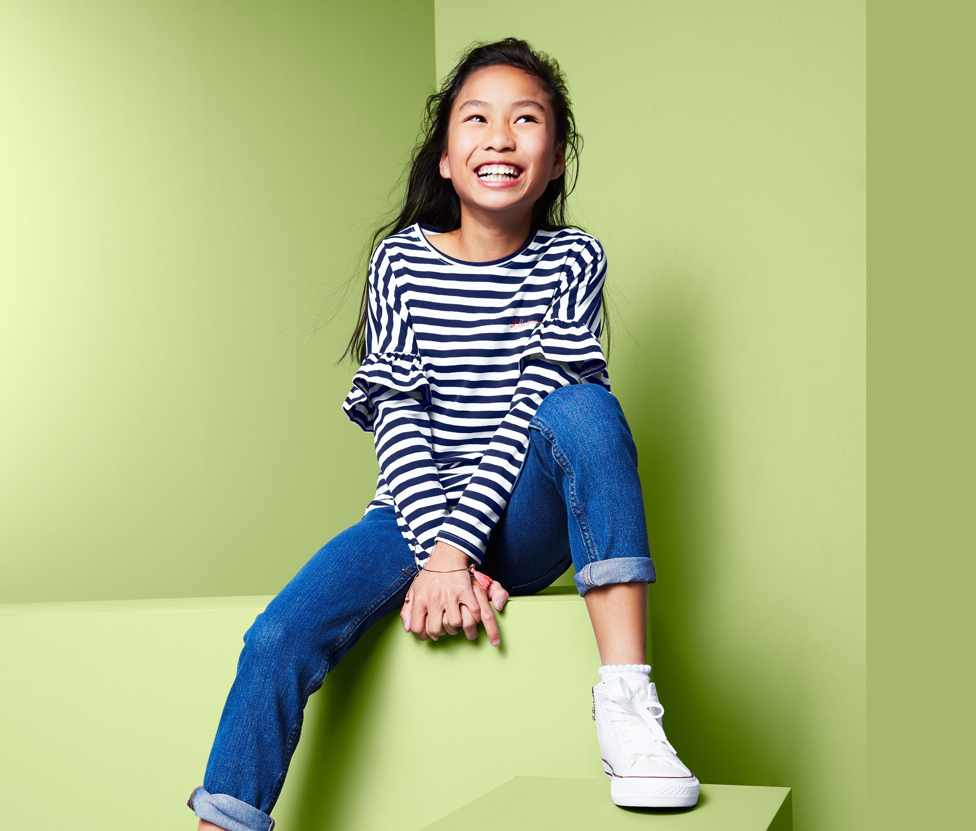 Lächelndes Mädchen sitzt auf grünen Blöcken und trägt ein Langarmshirt mit überschnittenen Ärmeln, Jeans und Turnschuhe.