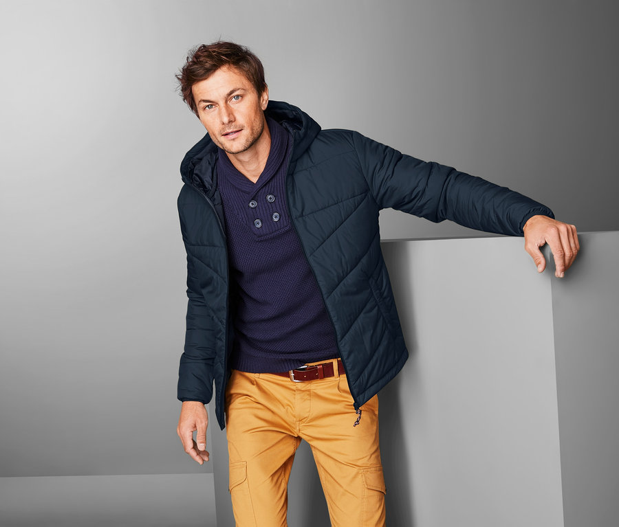 Mann posiert in Steppjacke, dunkelblauem Pullover und gelben Hosen.