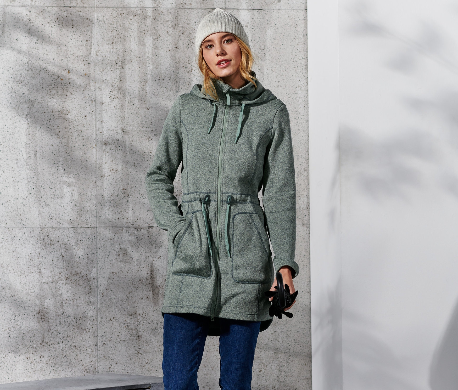 Frau posiert in Strickfleece-Mantel, Jeans, Stiefeln und Mütze.