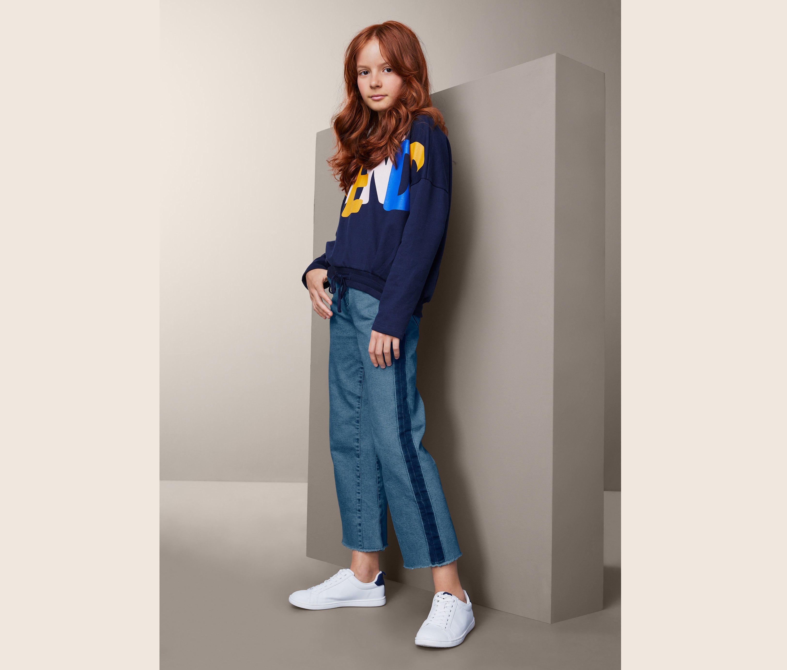 Ein junges Mädchen mit gewellten, roten Haaren posiert in einem dunkelblauen Sweatshirt, einer Jeans-Culotte mit Streifen und weißen Turnschuhen.