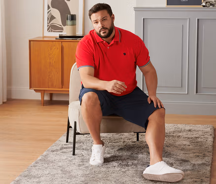 Mann sitzt auf einem Stuhl und trägt marineblaue Chino-Shorts, ein rotes Piqué-Poloshirt und Sneaker.