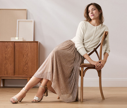 Frau sitzt auf Stuhl und trägt beigen Plisseerock.