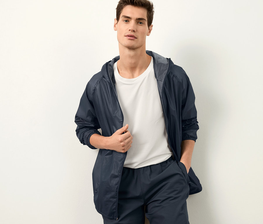 Mann posiert an einer Wand, trägt eine Windprotectionjacke und Shorts.