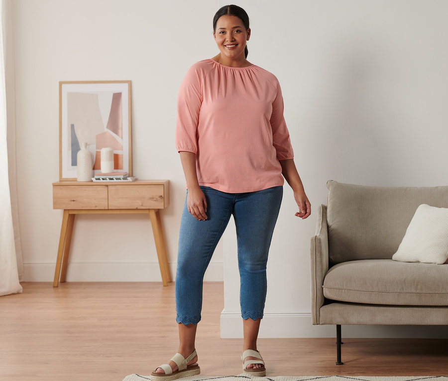 Eine Frau steht und trägt Plateau-Sandale aus Leder, 7/8-Treggings mit Lochstickerei und ein korallefarbenes Shirt mit 3/4-Arm.