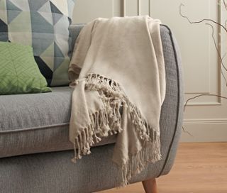 Eine beige Chenille-Wohndecke liegt auf einem grauen Sofa mit Kissen.