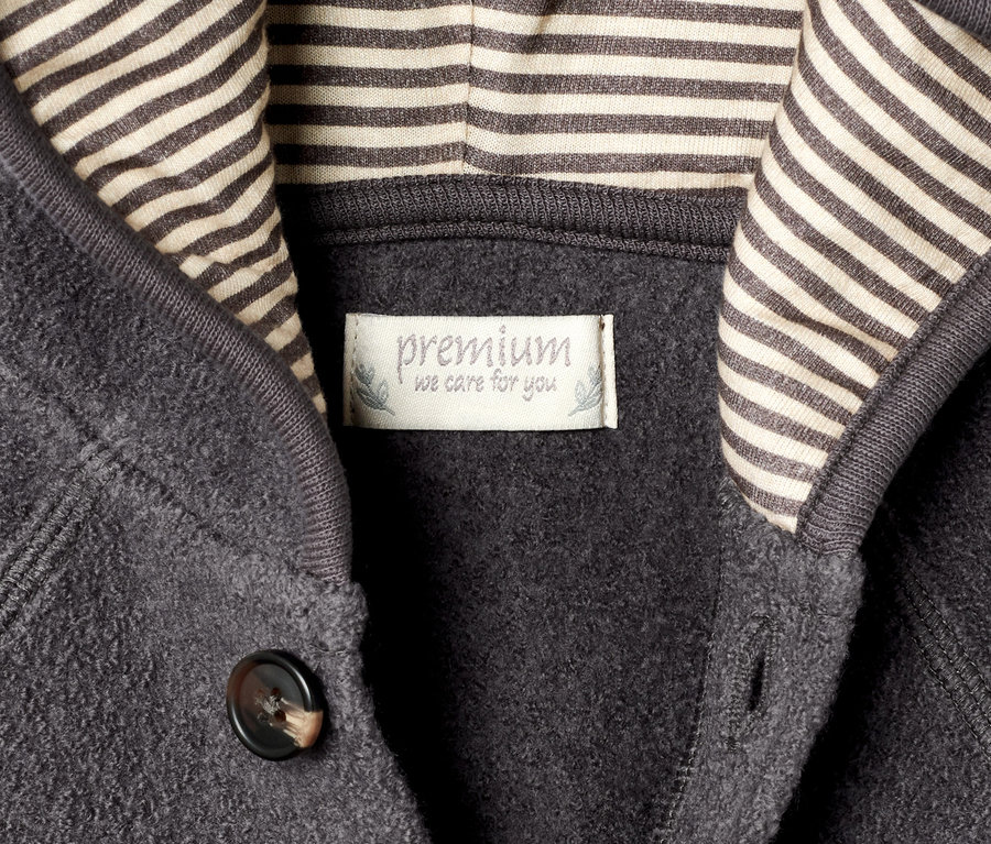 Detailansicht eines grauen Walkoveralls mit RWS-Wolle, einer Kapuze mit beigen Streifen und einem braunen Knopf.