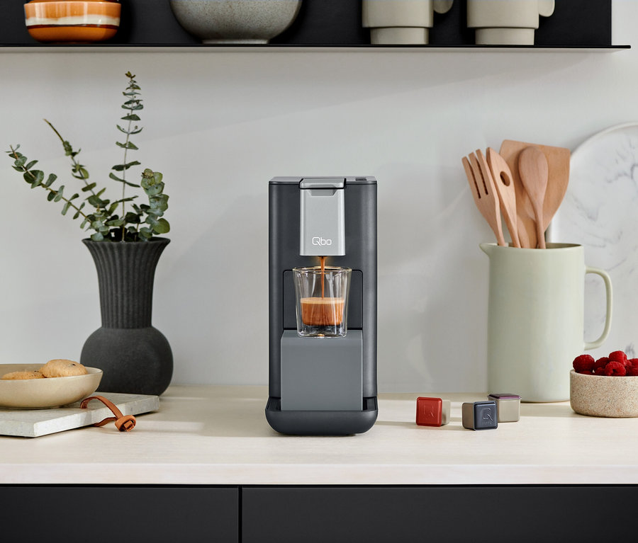 Qbo ESSENTIAL Dark Stone Kaffeemaschine befüllt ein Glas mit Kaffee. Kapseln liegen neben der Kaffeemaschine.