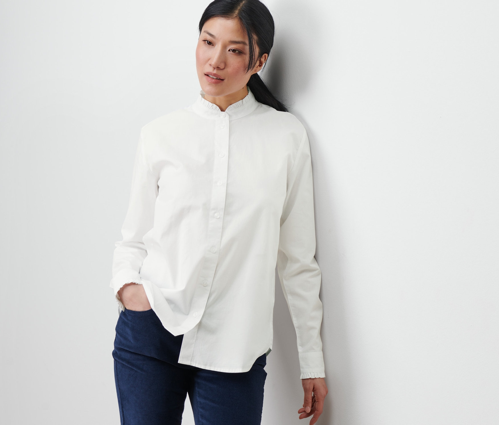 Eine Frau lehnt an einer Wand und trägt eine Bluse mit Rüschendetail und blaue Hosen.