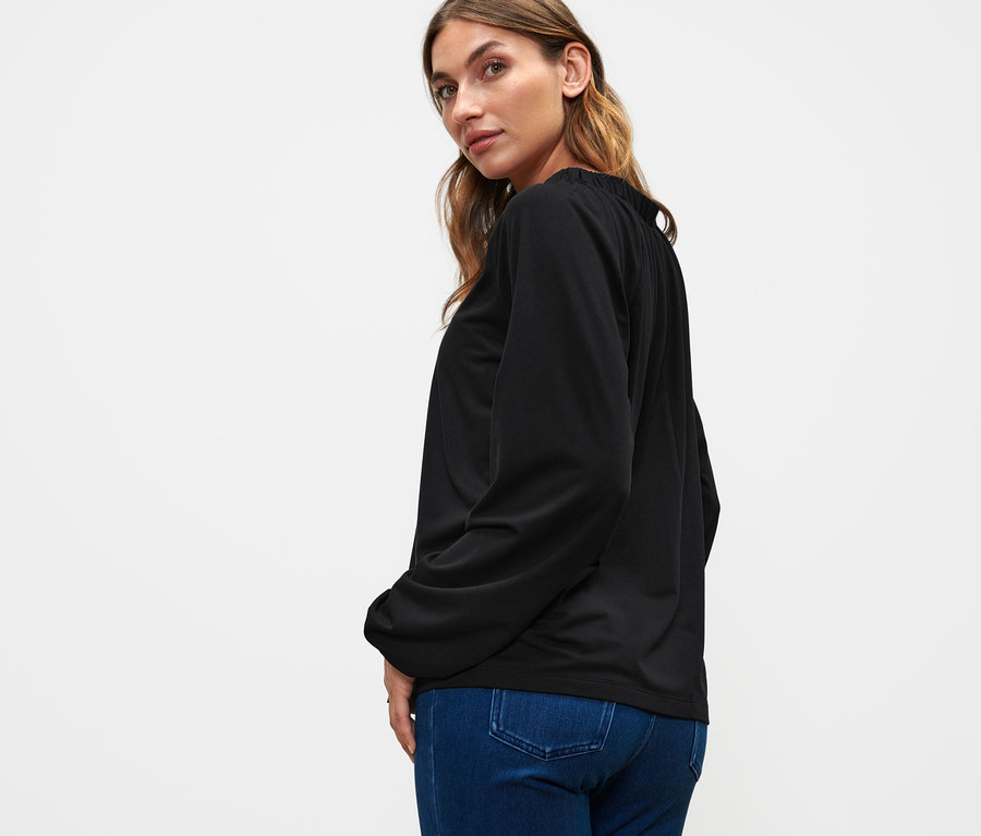 Frau blickt über die Schulter und trägt ein schwarzes Blusenshirt mit Raffung und Jeans.