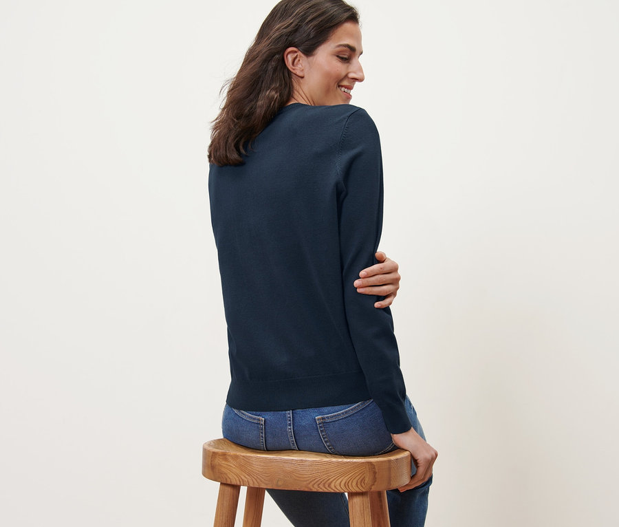 Frau sitzt seitlich auf einem Holzhocker und trägt einen marineblauen Feinstrick-Cardigan und Jeans.