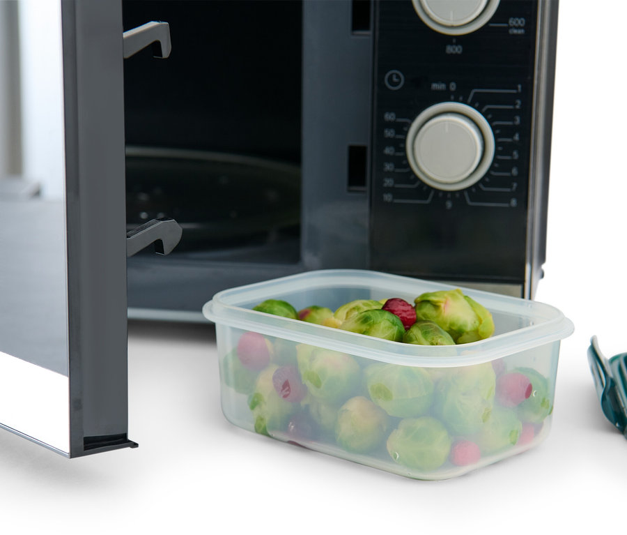Ein geöffneter Mikrowellenherd mit einer 3 Frischhaltedosen, die Rosenkohl und Himbeeren enthält. Neben der Dose liegt ein grüner Deckel.