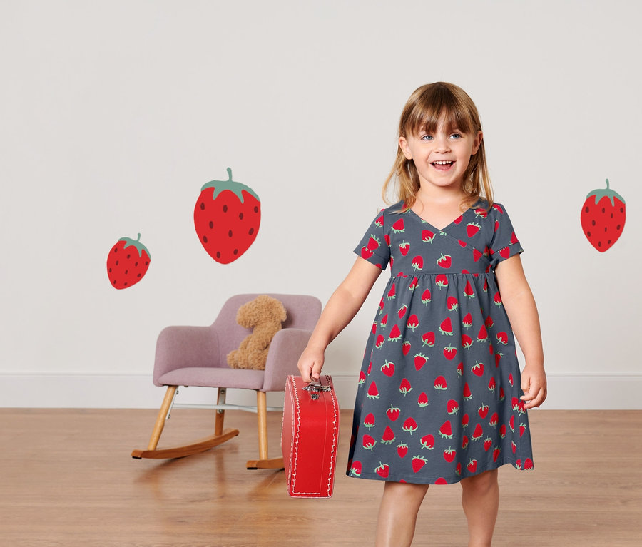 Lächelndes kleines Mädchen hält einen roten Koffer und trägt ein Kleinkinder-Jerseykleid mit Erdbeermuster.