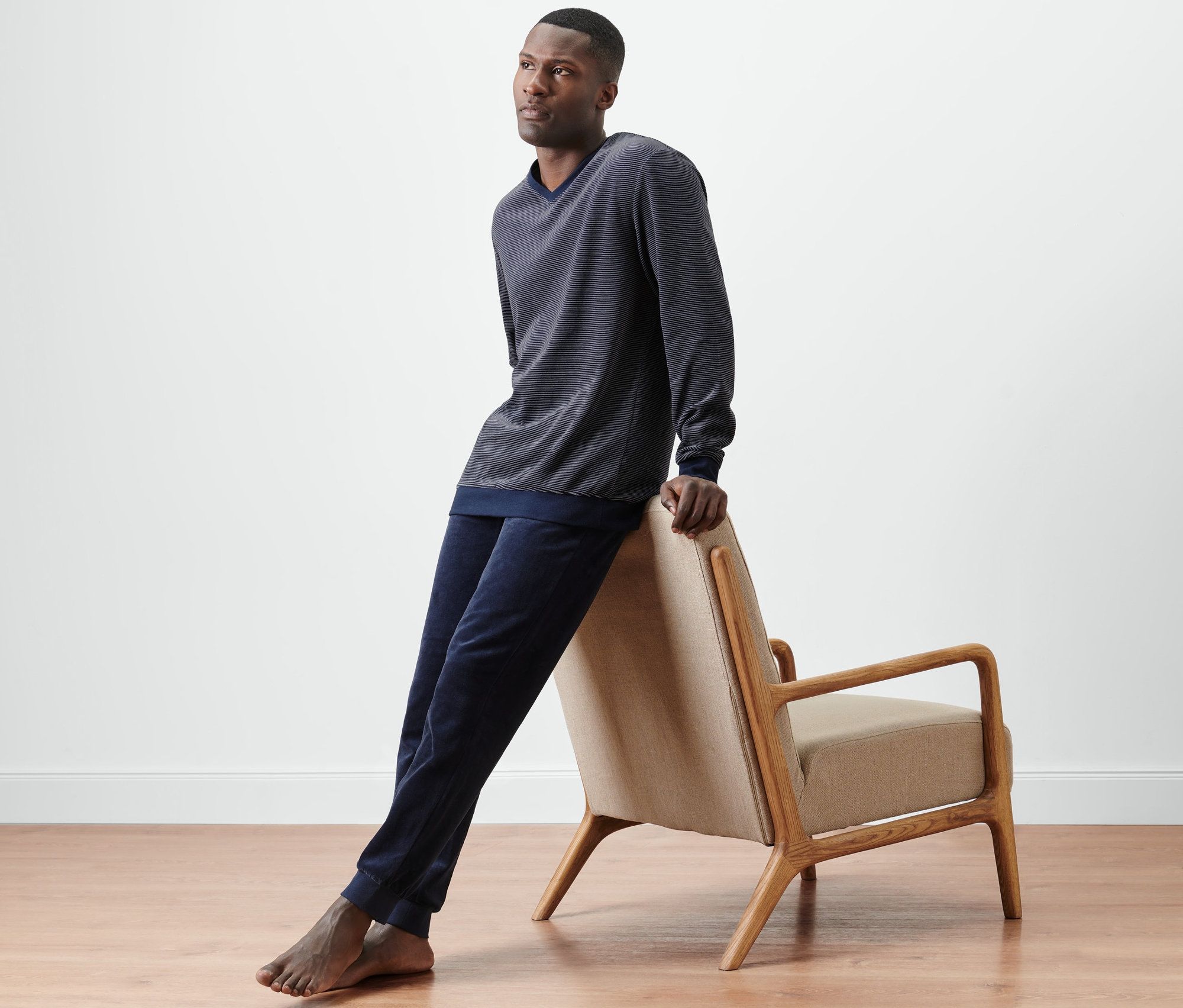 Mann lehnt an Sessel, trägt dunkelblau-grauen Nicki-Pyjama.