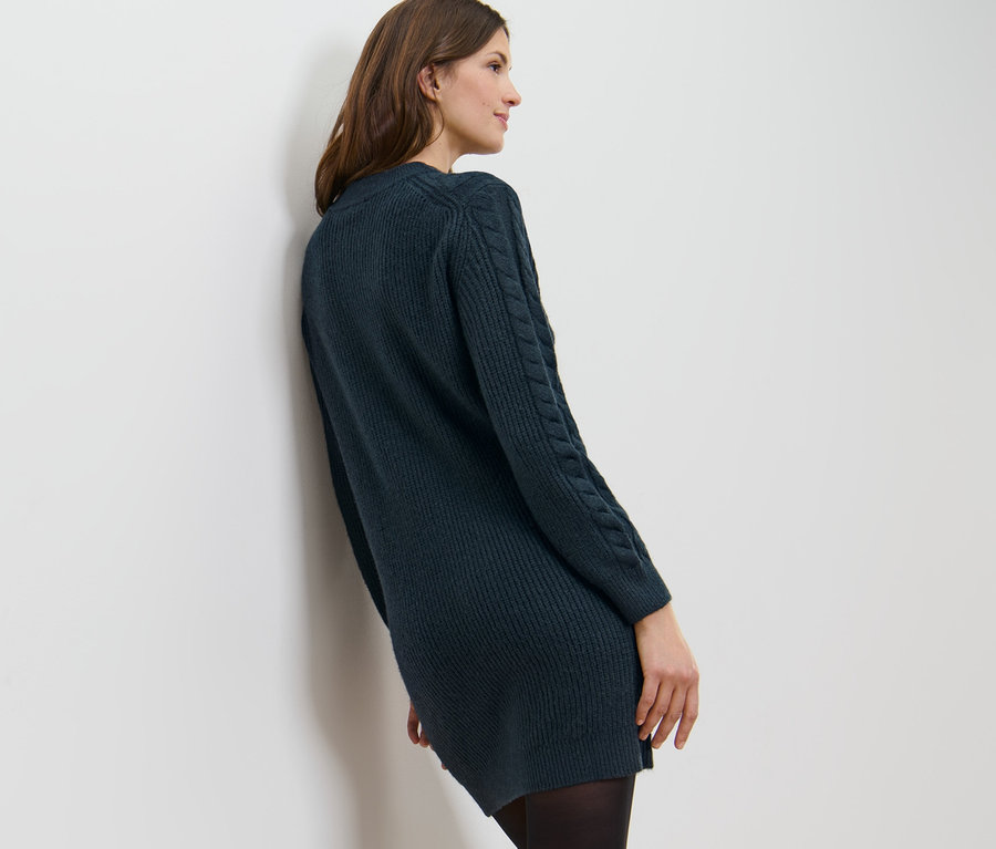Eine Frau lehnt an einer weißen Wand und trägt ein Strickkleid mit Zopfmuster.