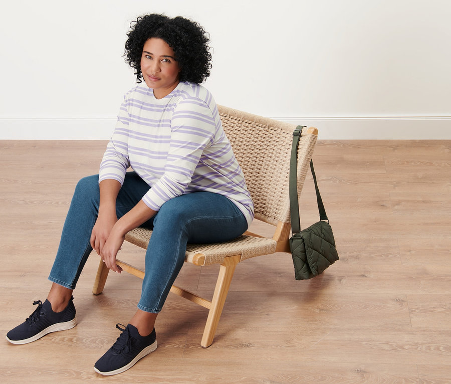 Eine Frau sitzt auf einem Korbstuhl und trägt Jeggings, ein lila-cremefarben gestreiftes Langarmshirt und eine gesteppte Umhängetasche.