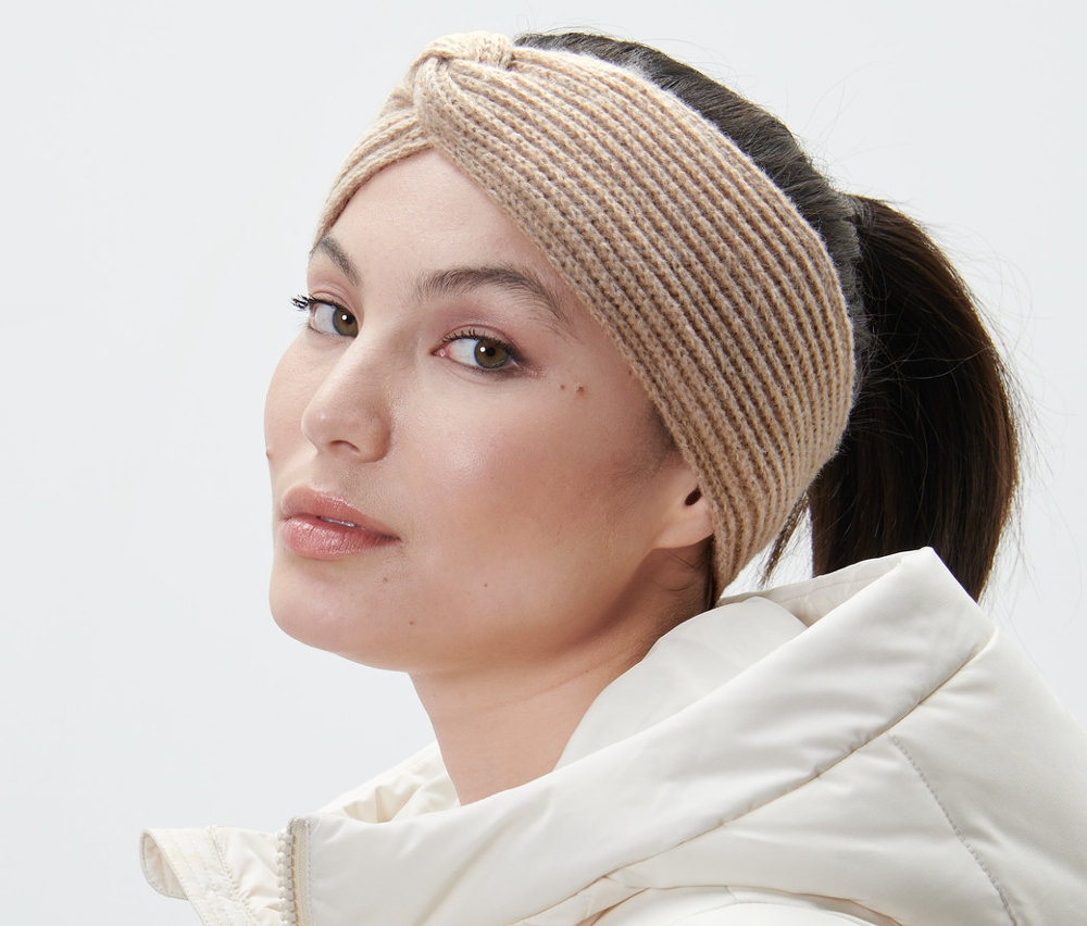 Frau mit sandfarbenem Strick-Stirnband und weißer Jacke blickt über die Schulter.
