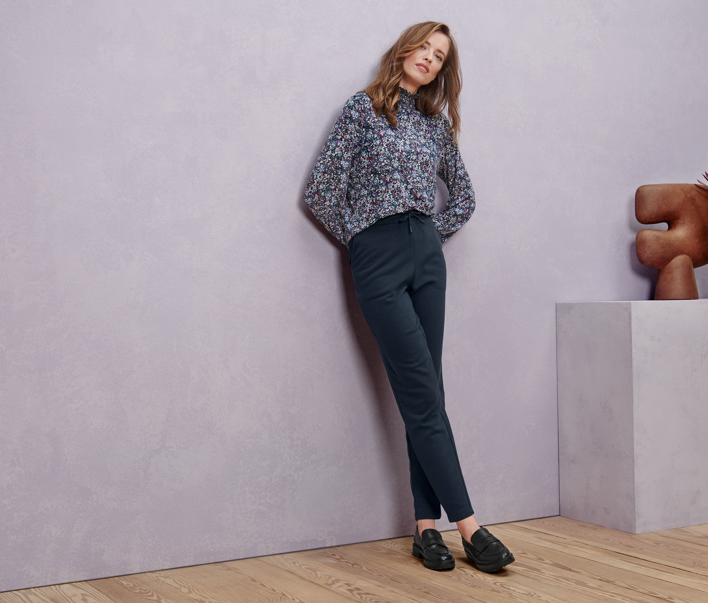 Eine Frau lehnt an einer Wand und trägt Leder-Loafer, ein Blusenshirt mit Stehkragen mit Millefleurs-Muster und eine Sweathose.