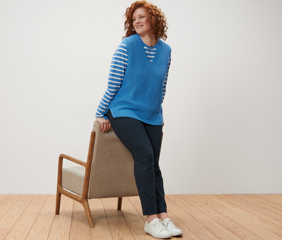 Eine Frau mit lockigem, rotem Haar lehnt an einem Stuhl und trägt ein gestreiftes Langarmshirt, einen Strick-Pullunder und eine dunkelblaue Bengalin-Hose.