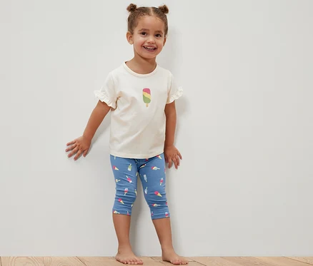 Ein lächelndes kleines Mädchen steht an einer weißen Wand und trägt eine blaue Dreiviertel-Leggings mit Eis am Stiel und ein weißes T-Shirt mit einem Eis am Stiel darauf. 2 ¾-Kinder-Leggings, blau.