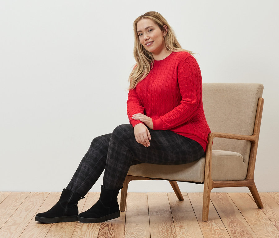 Frau sitzt auf einem Sessel und trägt einen roten Zopfstrickpullover und eine karierte Joggpants.