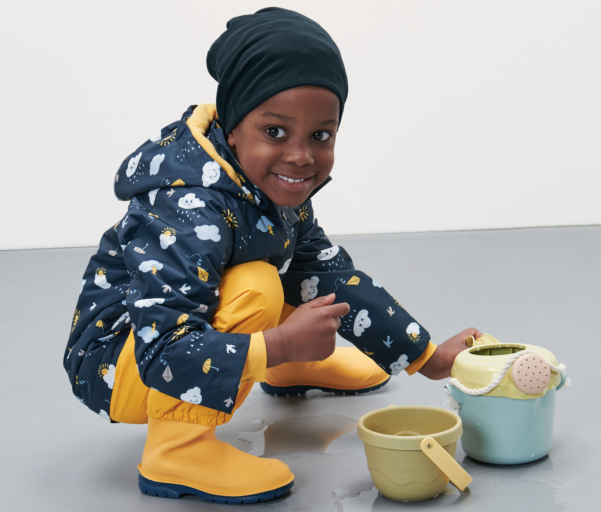 Ein Junge mit Kinder-Jerseymütze, bedruckter Jacke und gelben Gummistiefeln spielt mit Spielzeug auf dem Boden.