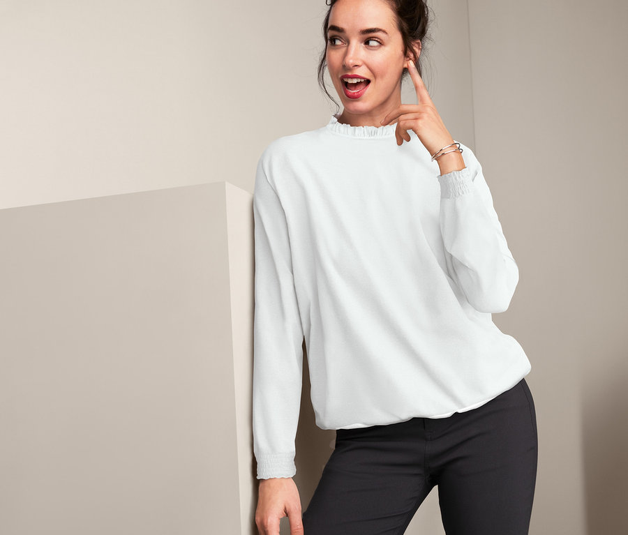 Frau lehnt an einer Wand und zeigt auf sich, sie trägt eine Bluse.