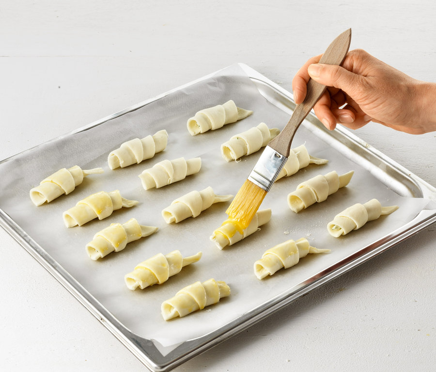 Eine Hand bestreicht mit einem Croissant-Roller rohe Croissants auf einem Backblech.
