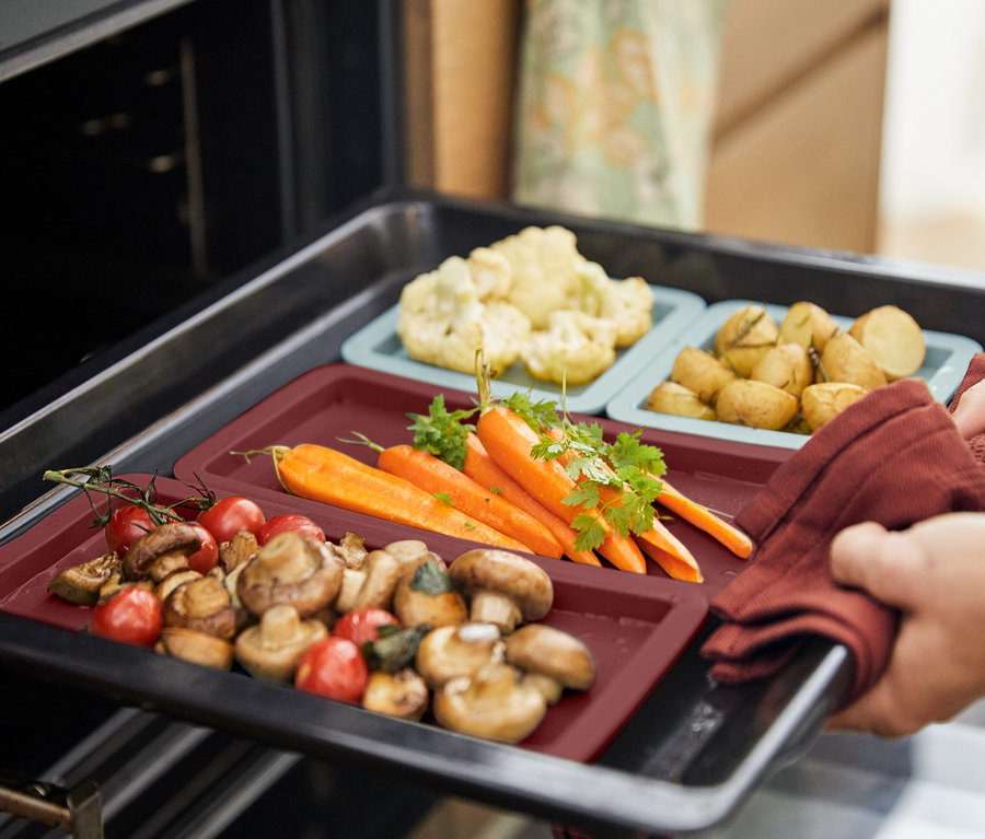 Eine Hand mit Handschuh nimmt ein Backblech mit 4 Backmatten aus dem Ofen, auf denen Gemüse liegt.