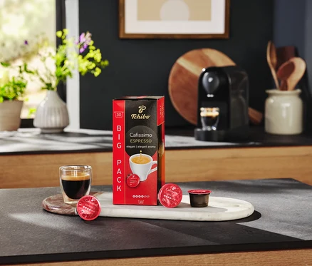 Auf einem Tisch stehen eine Packung Cafissimo Espresso elegant - 10 Kapseln, ein Glas Espresso und eine Kaffeemaschine.