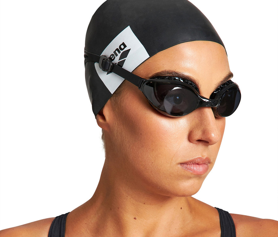 Frau trägt eine schwarze arena Badekappe, eine schwarze arena Air Soft Schwimmbrille und einen schwarzen Badeanzug.