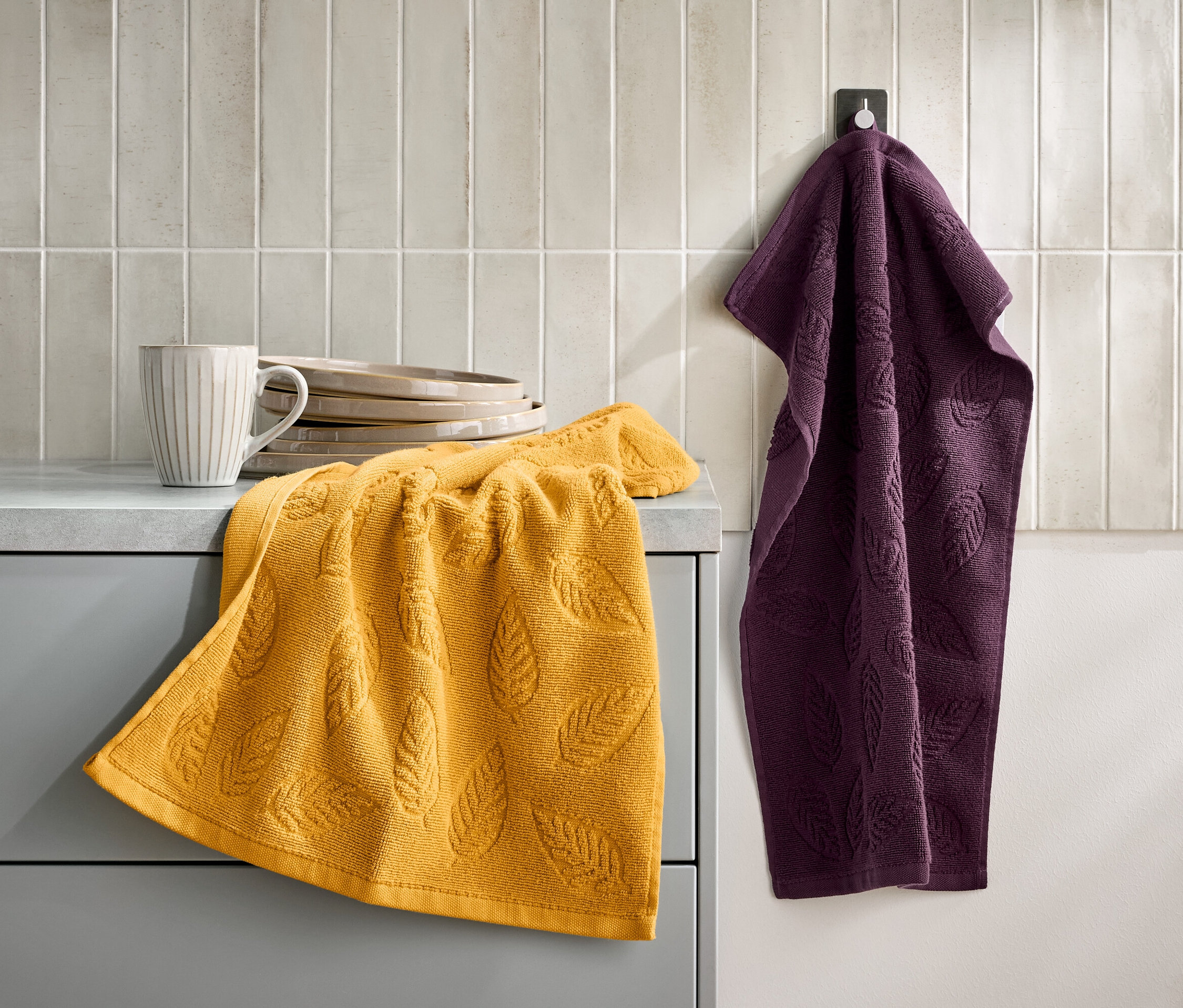 Ein gelbes Handtuch mit Blattmuster liegt auf einem grauen Schrank neben einer Tasse und Tellern. Ein lila Handtuch hängt an einem Haken an der Wand.