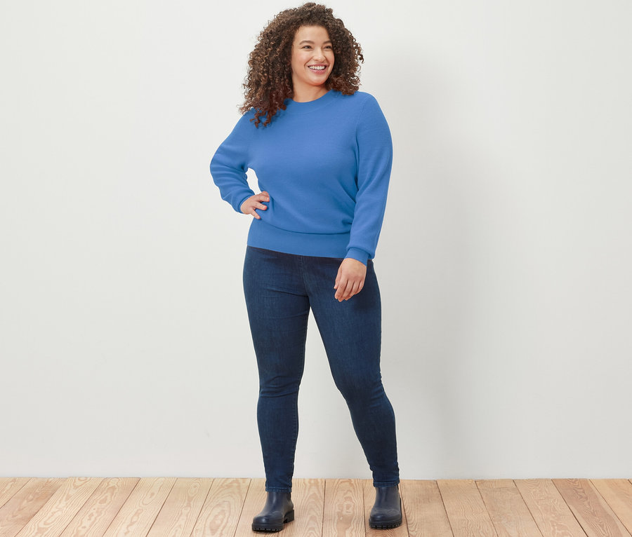 Frau posiert auf Holzboden in blauem Feinstrickpullover und Jeansleggings.