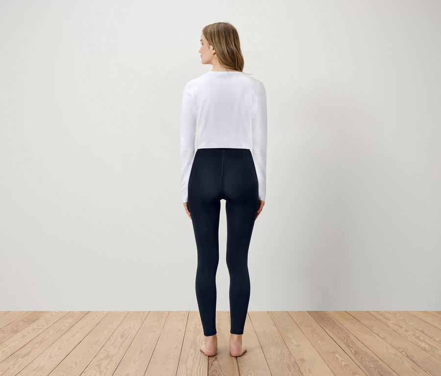 Frau steht mit dem Rücken zum Betrachter, trägt ein weißes Langarmshirt und dunkelblaue Leggings.