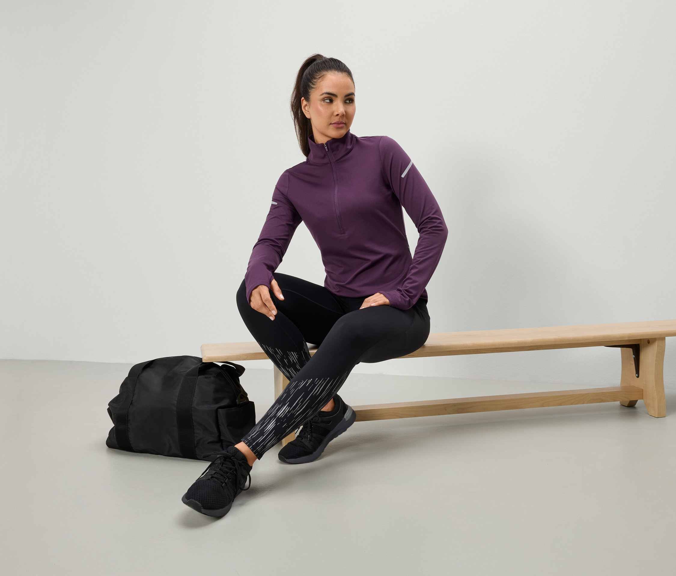 Frau sitzt in einem lila Sportoberteil und schwarzen Leggings auf einer Bank, neben ihr liegt eine schwarze Sporttasche.