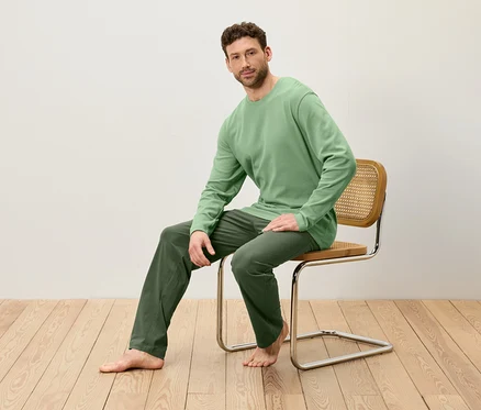 Mann sitzt auf einem Stuhl. Er trägt ein grünes Langarmshirt und eine grüne Hose.