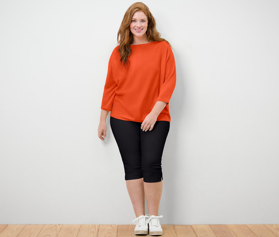 Eine Frau steht und posiert. Sie trägt einen orangefarbenen Feinstrickpullover und eine schwarze 3/4-Stretchhose.