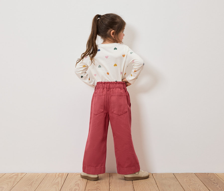 Mädchen mit Herzchenmuster-Shirt und roter Hose steht mit dem Rücken zur Kamera.