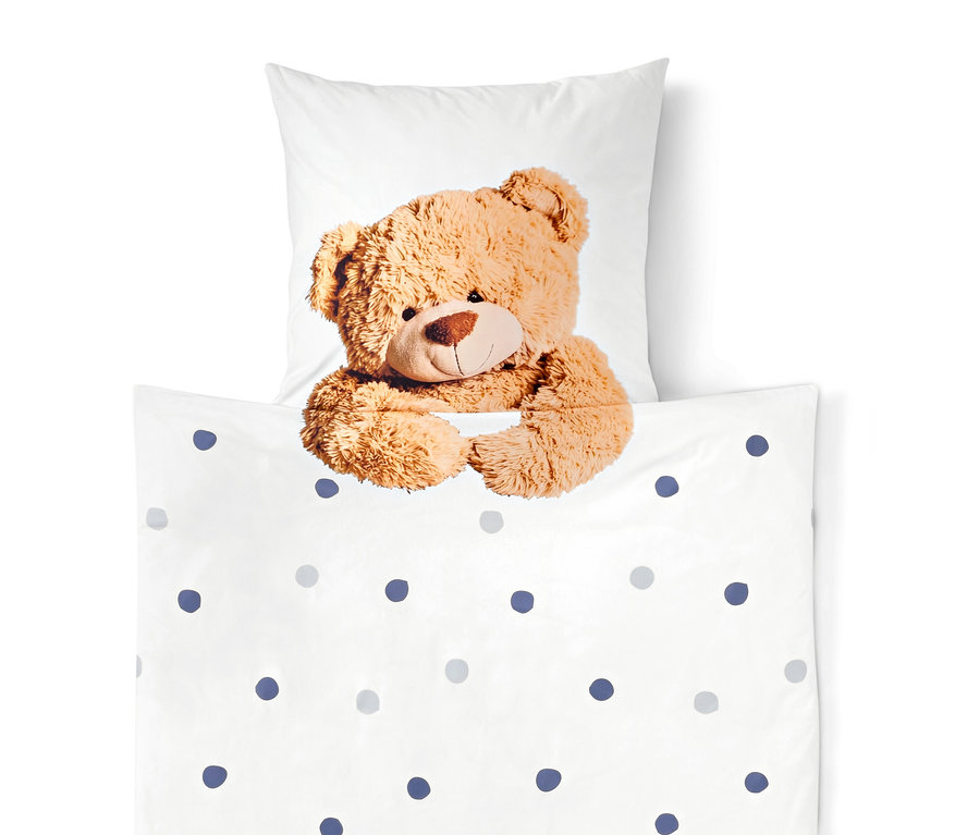 Ein brauner Teddybär lugt hinter einer weißen Decke mit blauen und grauen Punkten hervor, ein weißes Kissen im Hintergrund.