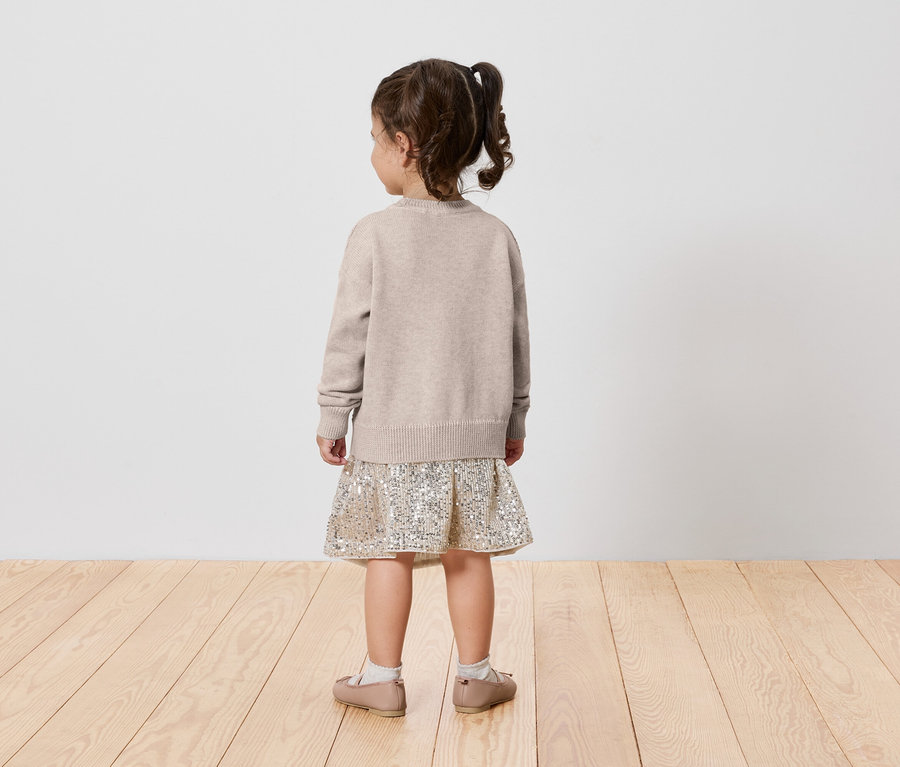 Ein kleines Mädchen mit einem beigen Pullover und einem gemusterten Rock steht mit dem Rücken zur Kamera.