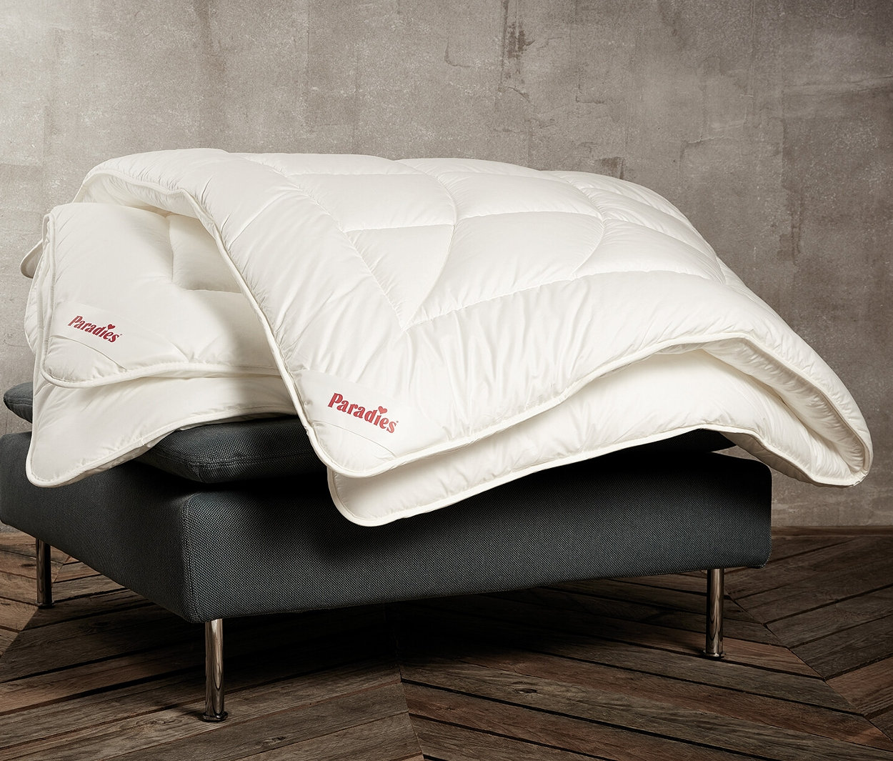 Auf einem grauen Sessel liegen eine normalgroße und eine übergroße, weiße Paradies Ganzjahresdecke »ElbTraum«.