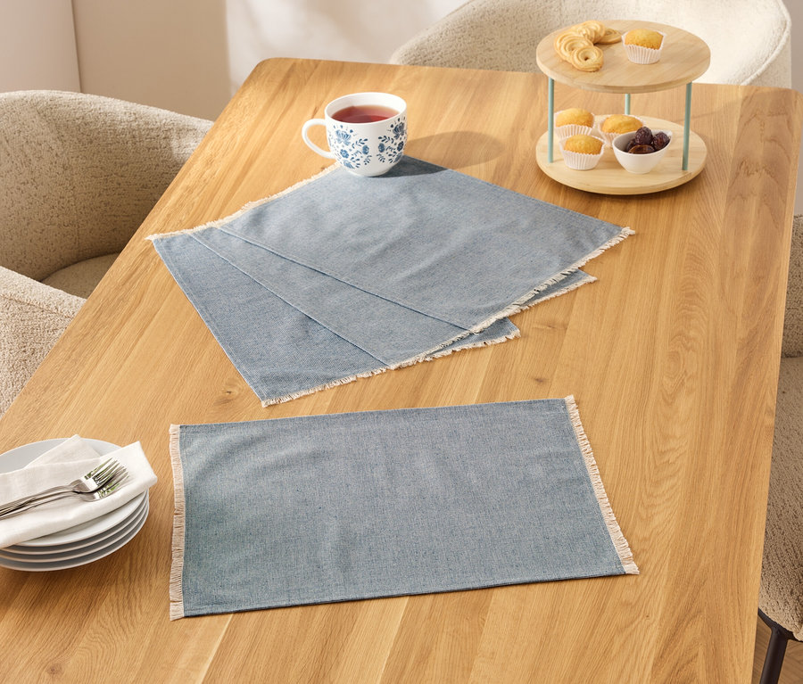 Zwei blaue Tischsets mit weißem Rand auf einem Holztisch, daneben eine Tasse, Teller und eine Etagere mit Essen.